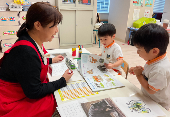 先生が子どもたちと一緒に、テーブルに広げた教材を使って学習している様子。子どもたちは興味深そうに本を見ています。